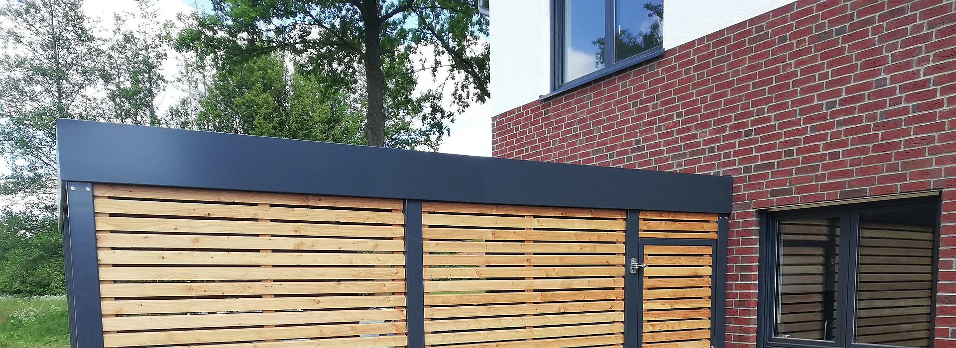 anthrazit farbenes Carport aus Metall und Holz in Einfahrt mit angrenzendem Holzschuppen, Seiteneingang